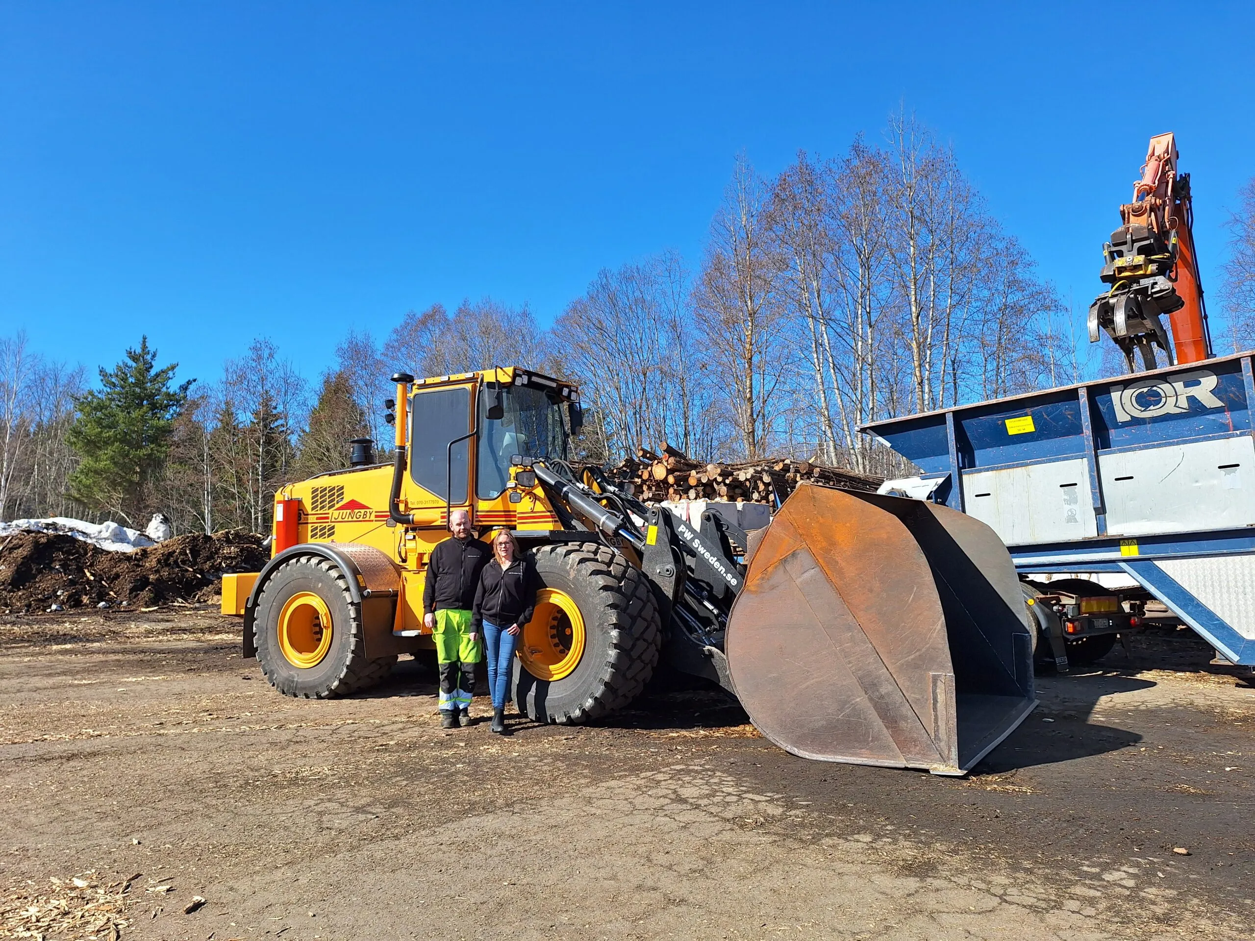 Två personer framför en gul hjullastare på industriplanen